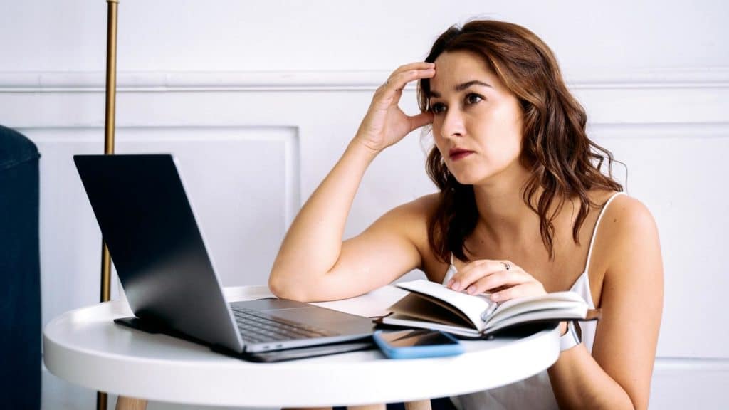 Frau sitzt auf dem Boden vor kleinem Tisch. vor ihr ein Laptop und ein Notizbuch. Sie hält sich den Kopf und wirkt erschöpft.